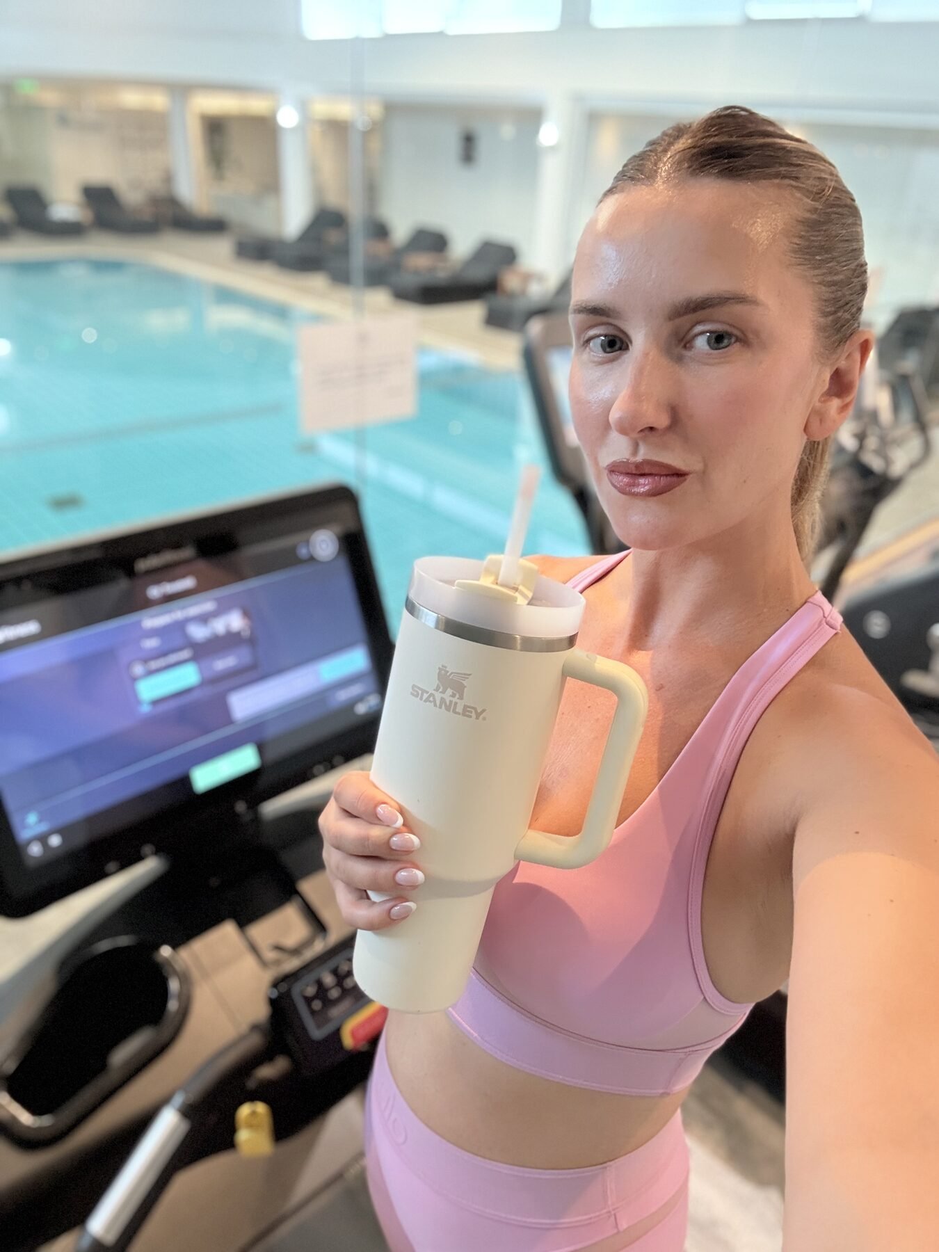 Woman holding a cream Stanley Quencher tumbler while exercising on a treadmill beside an indoor swimming pool.